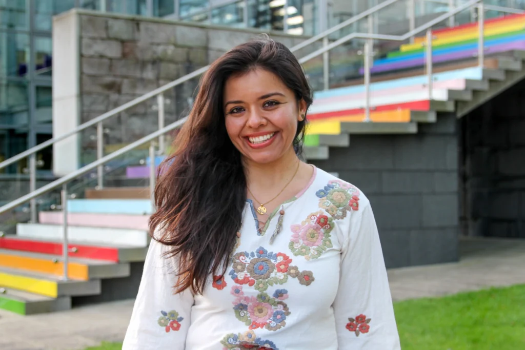 Sandra Martinez, owner of The Wee Web Studio, smiling in front of the rainbow-coloured outdoor stairs in Aberdeen. She is wearing a white top with colourful floral embroidery.